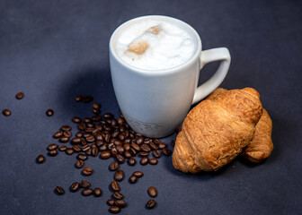 Beautiful latte macchiato with milk foam in a white cup with croissants and coffee beans nearby....