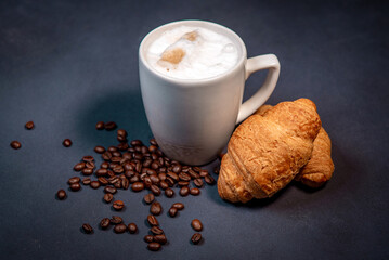 Beautiful latte macchiato with milk foam in a white cup with croissants and coffee beans nearby. Dark blue background. A cup of coffee with milk. Hot latte or cappuccino on milk with copy space