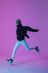 Fototapeta na wymiar Young stylish man in modern street style outfit isolated on gradient background in neon light. African-american fashionable model in look book, musician performing.
