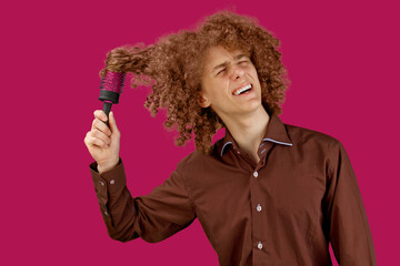 A long-haired curly-haired guy in a brown shirt on a red background uses a wooden comb. Emotions before a haircut in a hairdresser. Pain from combing
