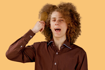A long-haired curly-haired guy in a brown shirt on an orange background uses a wooden comb....