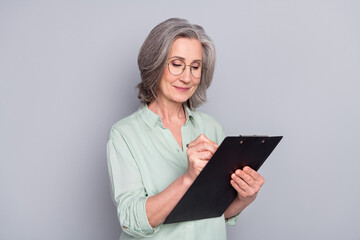 Photo of happy focused peaceful concentrated woman in glasses writing in checklist isolated on grey color background