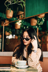 Korean young girl drinks coffee in black glasses, in a beige blouse in a cafe