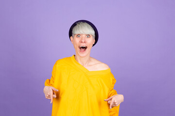 Stylish european woman on purple background pointing down with fingers showing advertisement, surprised face and open mouth