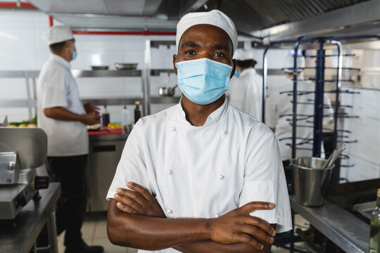 Portrait of african american male professional chef wearing face mask with colleagues in background