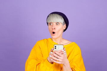 Stylish european woman on purple background with mobile phone surprised amazed shocked open mouth