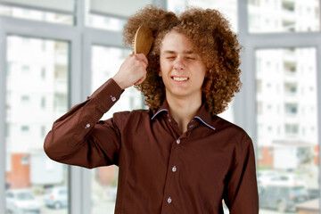 A long-haired curly-haired guy in a brown shirtuses a wooden comb. Emotions before a haircut in a...