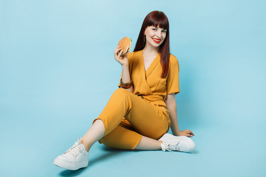 Diet and jank food concept. Portrait of young red haired beautiful woman holding fresh burger and smiling to camera, sitting on the floor over isolated blue background