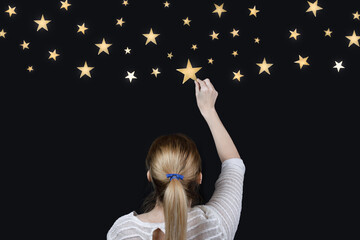 Back view of blonde girl with ponytail stretching her arm to catch bright stars in the night sky....