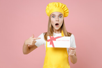 Teen girl chef cook confectioner housewife mother's helper baker in yellow apron white tshirt cap point index finger on gift voucher flyer mock up isolated on pink background studio Food cake concept