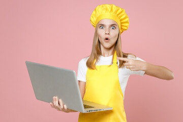 Teen shocked girl chef cook confectioner housewife baker wearing yellow apron white t-shirt cap point index finger on laptop pc computer isolated on pink background studio portrait Food cake concept.