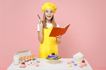 Teen proactive fun girl chef cook confectioner baker in yellow apron cap at table writing recipe idea in notebook isolated on pastel pink background studio Mousse cake food process workshop concept