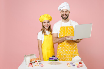 Teen fun happy girl dad father chef cook confectioner baker in yellow apron cap at table read look for recipe on pc laptop isolated on pink background Mousse cake food workshop master class process.