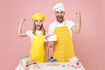 Teen fun girl dad chef cook confectioner baker in yellow apron cap at table hold gift voucher flyer mockup do winner gesture isolated on pink background Mousse cake food workshop master class process