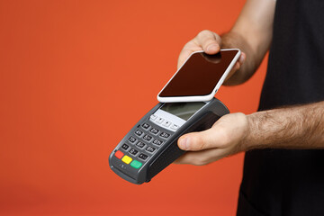 Close up cropped photo shot young man barista barman employee in apron white t-shirt work in coffee shop use wireless bank payment terminal credit payment mobile phone isolated on orange background