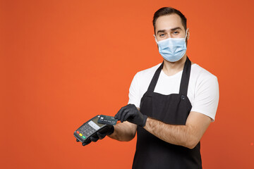 Young man barista barman employee in apron white tshirt face mask coronavirus covid19 quarantine work in shop uses wireless bank payment terminal process acquire payment isolated on orange background