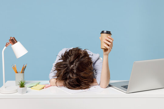 Young tired exhausted secretary employee business woman in casual shirt sit work sleep laid her head down on white office desk with pc laptop hold cup coffee isolated on pastel blue background studio.