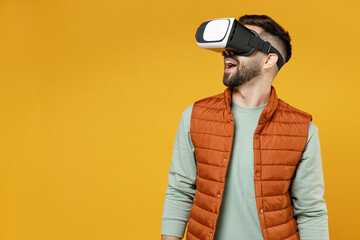 Young smiling happy man 20s wear orange vest mint sweatshirt glasses watching in vr headset pc gadget isolated on yellow color background background studio portrait. Entertainment lifestyle concept