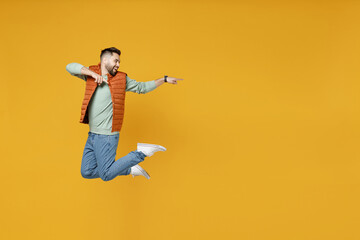 Full length side view young smiling happy fun caucasian man 20s wear orange vest mint sweatshirt jump high point index finger aside on copy space workspace area isolated on yellow background studio.