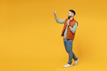 Fototapeta premium Full length young smiling friendly man in orange vest mint sweatshirt glasses do selfie shot on mobile phone show victory v-sign gesture isolated on yellow background studio. People lifestyle concept.