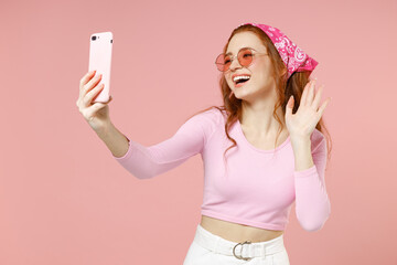 Fun young cheerful friendly happy woman 20s wear rose clothes bandana glasses doing selfie shot on mobile phone waving hand isolated on pastel pink background studio portrait People lifestyle concept.