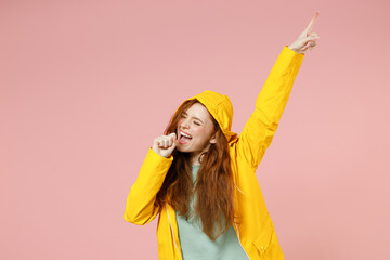 Redhead young fun woman 20s in yellow waterproof raincoat outerwear sing song in microphone point finger up isolated on pastel pink color background studio. Outdoors lifestyle wet fall season concept.