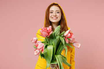 Redhead young smiling happy woman in yellow waterproof hood raincoat outerwear giving tulips flower bouquet isolated on pastel pink background studio Lifestyle spring weather blossom season concept.