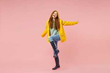 Full length redhead happy young ginger woman in yellow waterproof raincoat hood outerwear raised up leg isolated on pastel pink background studio Outdoors lifestyle wet fall weather season concept