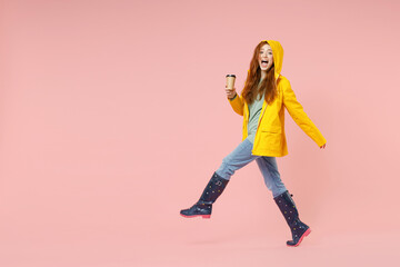 Fototapeta na wymiar Full length side view redhead young woman in yellow waterproof hood raincoat outerwear holding paper cup of coffee drink hot tea walk isolated on pastel pink background. Lifestyle fall season concept.
