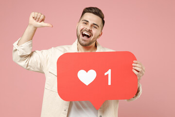 Young fun cheerful caucasian man 20s wear jacket white t-shirt huge like sign from social network heart form point index finger on himself isolated on pastel pink color background studio portrait