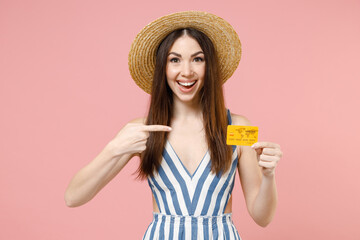 Young fun caucasian rich smiling happy woman 20s in summer clothes striped dress straw hat put index finger on credit bank card looking camera isolated on pastel pink color background studio portrait