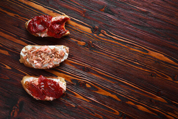 three bruschetta sandwiches on baguette bread with cottage cheese, sun-dried tomatoes and red fish on a dark wooden background, top view, copy space. Traditional italian snack for wine - grilled tapas