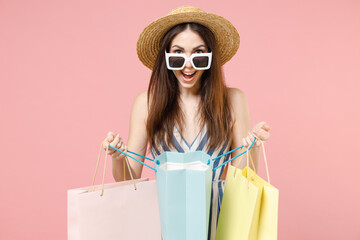 Young overjoyed caucasian woman wear summer clothes striped dress straw glasses hold package bags with purchases after shopping touch hat look camera isolated on pastel pink color background studio.