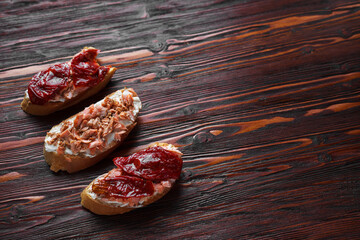 three bruschetta sandwiches on baguette bread with cottage cheese, sun-dried tomatoes and red fish on a dark wooden background, top view, copy space. Traditional italian snack for wine - grilled tapas