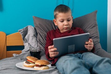 young boy eating unhealthy food and using digital tablet while sitting in his room