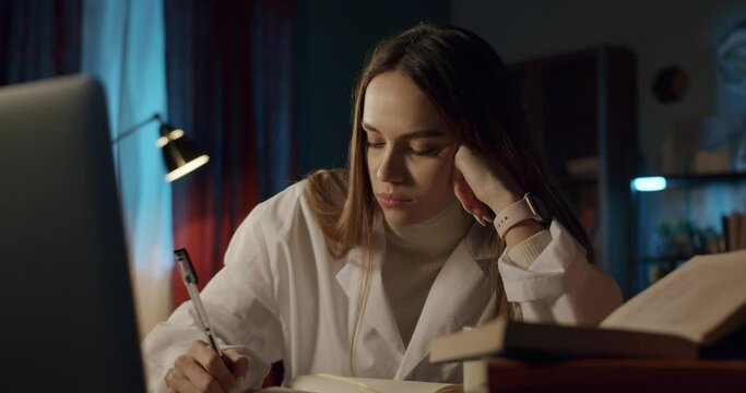 Tired young woman wearing medical uniform gown, female intern working at night with a computer. Doctor nurse using laptop and video call, writing notes, overtime at the workplace, headache.