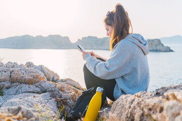 Caucasian woman using her smartphone on a cliff by the sea at the Malgrats Islands in Santa Ponça. Palma de Mallorca, Spain