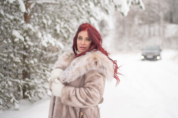 young beautiful woman with red long hair stands in the winter park forest snow in a fur coat