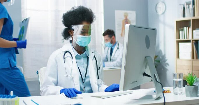 Portrait of beautiful busy African American young woman physician in medical mask and face shield typing on computer searching internet sitting in cabinet in medical center. Medic concept