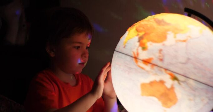 Boy Playing With Globe at Night. Child Sits on Bed in Evening Light Dreaming Vacation. Little Child Looking at Illuminated Globe, Exploring World, Learning. Dreaming About Future Save of Our Planet.