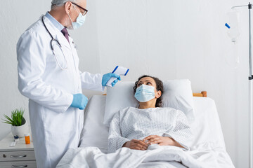 mature doctor in medical mask measuring temperature of sick african american woman