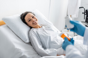 smiling african american woman lying in hospital bed near doctor with medication on blurred foreground