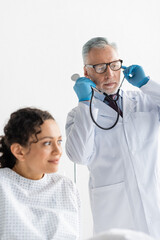 doctor in latex gloves putting on stethoscope near african american woman on blurred foreground