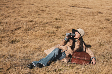 Woman traveler with hat and suitcase is lying down in a golden grass field and is drinking water from her thermos bottle.