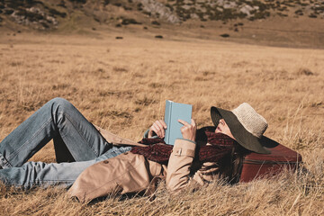 Woman traveler with hat is lying down in a golden grass field and is reading a book.