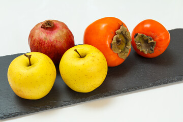 Apples, pomegranate and ripe persimmon fruit on the black stone board.
