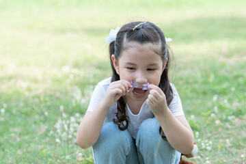 Little cute Caucasian kid girl with twin tails hair smelling small purple flowers in her hands and sitting on green grass at park on summer