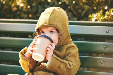 Funny toddler kid resting on bench, holding paper cup of take away coffee