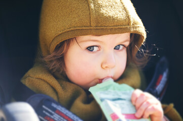 Close up portrait of sweet toddler kid eating fruit puree from plastic doy pack, sitting in stroller, outdoor snack time - 429422734