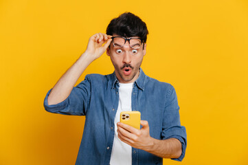 Fototapeta Amazed shocked caucasian guy holding smartphone in his hand, looking at the phone in surprise with his glasses raised, stunned facial expression, stands on isolated orange background obraz